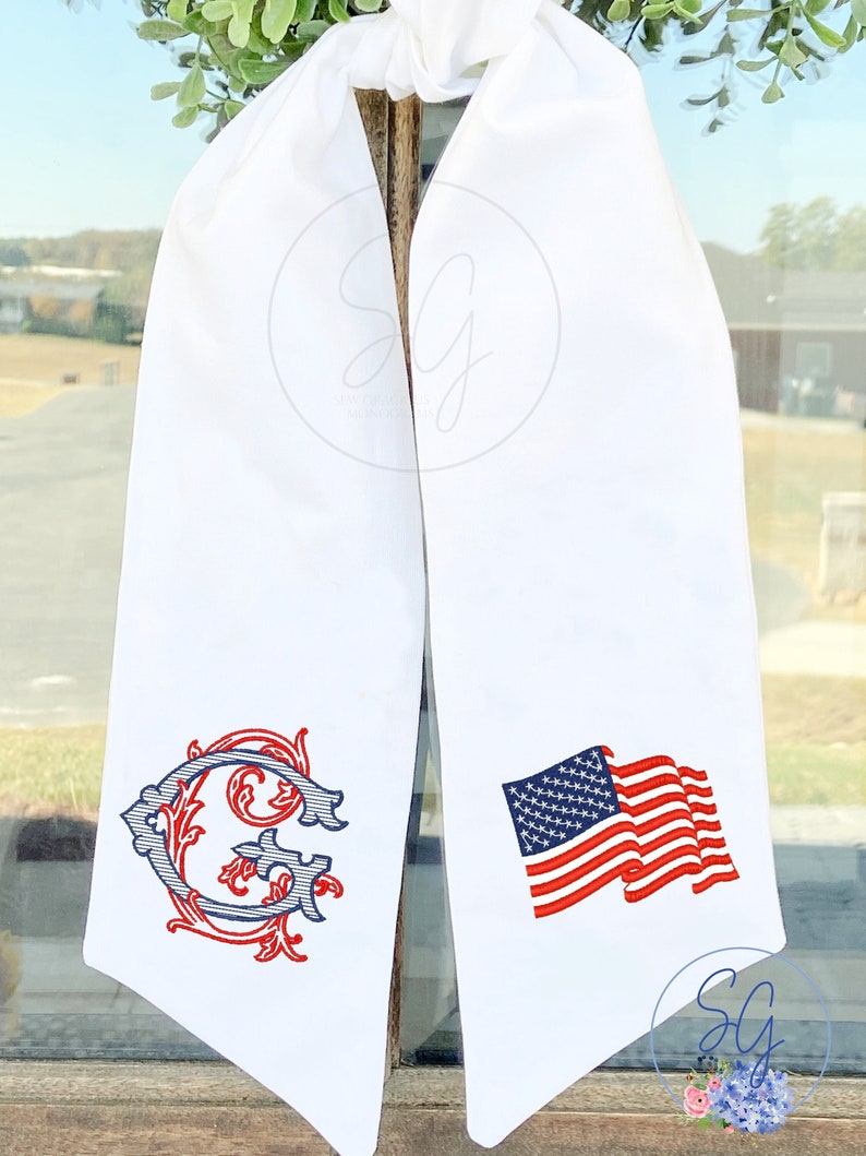 May include: White fabric sash with a red, white, and blue embroidered American flag on one side and a red and blue embroidered letter G with floral embellishments on the other side.