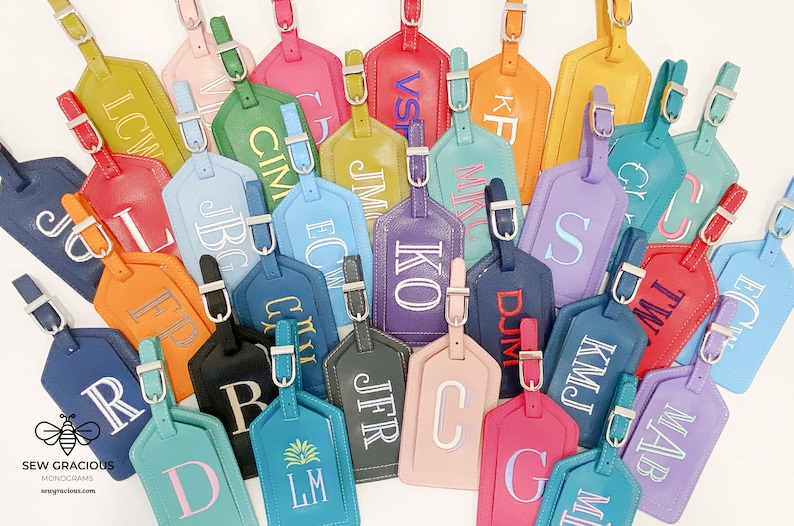 May include: A collection of colourful leather luggage tags with personalised initials in white embroidery. The tags are arranged in a random pattern on a white surface.