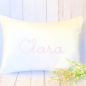 May include: A white pillow with the name "Clara" embroidered in pink thread. The pillow is on a white wooden surface with a bouquet of white and pink flowers.