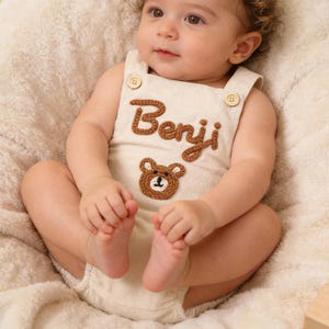 Puede incluir: Un pelele para bebé de color crema con el nombre Benji bordado en hilo marrón y una pequeña aplicación de una cara de oso marrón. La prenda tiene dos botones de madera clara en los tirantes y un diseño sencillo sin mangas.