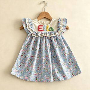 May include: A light blue floral print dress with the name "Ella" embroidered in colorful thread. The dress features ruffled sleeves and a cream-colored collar. The dress is hanging on a wooden hanger.