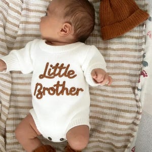 Może przedstawiać: Białe dzianinowe body z krótkim rękawem z napisem Little Brother wyhaftowanym brązową nicią na piersi. Ubranko zestawiono z pasującą brązową dzianinową czapką i brązowymi bucikami, umieszczonymi na tkaninie w paski.
