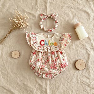 Puede incluir: Un conjunto para beb&eacute; que incluye un pelele con estampado floral en rosa y rojo, bordado con el nombre "Chloe" en hilo multicolor. Incluye una diadema a juego y un biber&oacute;n. El pelele tiene mangas y ribetes con volantes.