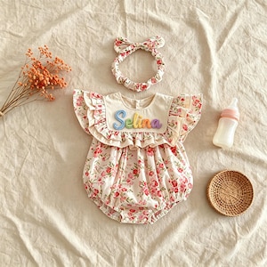 Puede incluir: Un conjunto floral para beb&eacute; que incluye un pelele y una diadema a juego. El pelele presenta un corpi&ntilde;o color crema con el nombre "Selina" bordado en hilo arco&iacute;ris. El pelele y la diadema tienen un estampado floral rosa. Tambi&eacute;n hay un biber&oacute;n y una cesta.