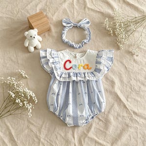 Puede incluir: Conjunto de pelele y diadema a juego para beb&eacute;. El pelele tiene un cuello blanco bordado con el nombre "Cora" en hilo de colores. El pelele es de tela a rayas azules y blancas con lazos. Un peque&ntilde;o osito de peluche blanco y un bloque de madera tambi&eacute;n est&aacute;n en la imagen.