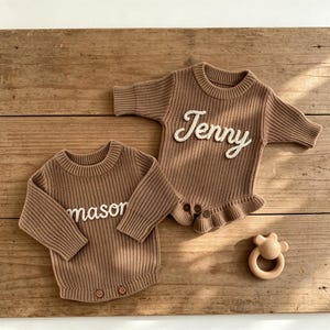 May include: Two brown knit baby rompers with ribbed textures and long sleeves. One romper features the white embroidered name Jenny, and the other features the white embroidered name mason. Both include two small brown buttons at the bottom.