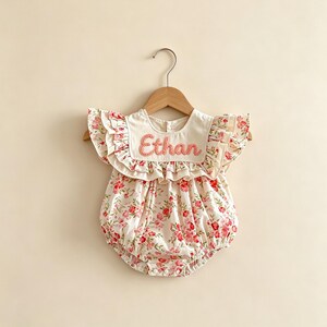 Puede incluir: Un pelele con estampado floral con el nombre "Ethan" bordado en rosa. El pelele tiene un cuello blanco y mangas con volantes. La tela es blanca con un estampado de flores rojas y rosas.