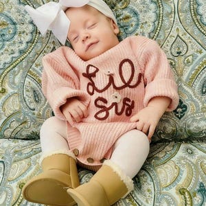 May include: A baby wearing a light pink ribbed knit long-sleeve romper with the text "Lil Sis" embroidered in dark brown, a white headband with a large bow, white tights, and tan ankle boots with a cream-colored faux fur lining at the top.