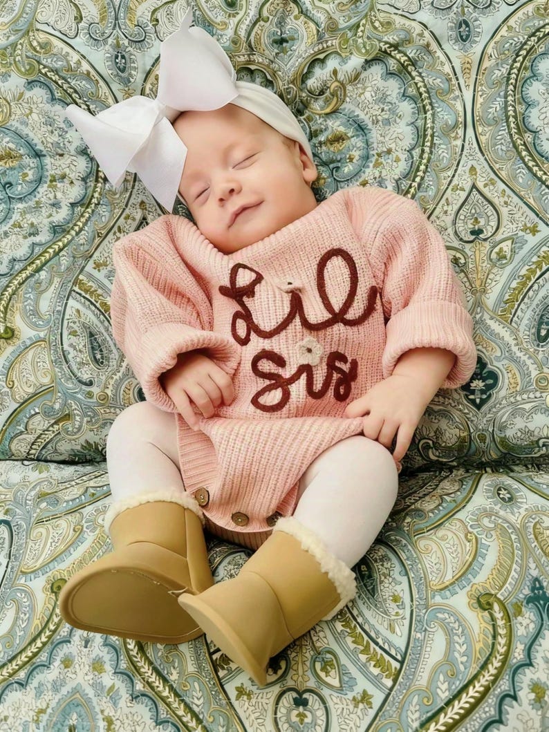 May include: A baby wearing a light pink ribbed knit long-sleeve romper with the text "Lil Sis" embroidered in dark brown, a white headband with a large bow, white tights, and tan ankle boots with a cream-colored faux fur lining at the top.