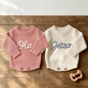 Puede incluir: Dos jerséis de punto acanalado de manga larga, uno en rosa empolvado con el nombre Ola bordado en blanco y otro en crema con el nombre Jetson bordado en azul claro. Ambos jerséis tienen cuello redondo y dos botones de madera en el dobladillo.