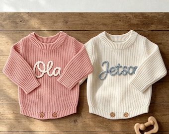 May include: Two long-sleeved ribbed knit sweaters, one in dusty pink with the white embroidered name Ola and one in cream with the light blue embroidered name Jetson. Both sweaters feature a crew neckline and two wooden buttons at the bottom hem.