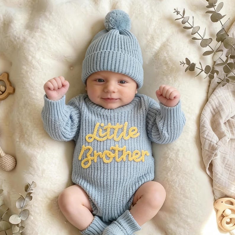 May include: A light blue knitted long-sleeve romper with a matching beanie featuring a pom-pom. The romper has the text Little Brother embroidered in yellow cursive script across the chest. The set includes matching light blue knitted booties.