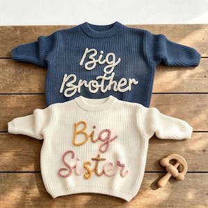 May include: A dark blue knit sweater with the white text Big Brother and a cream knit sweater with the multicolored text Big Sister, featuring a wooden rattle toy. Both sweaters have long sleeves, ribbed crew necklines, and cuffs.