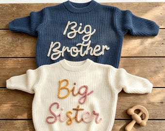 May include: A dark blue knit sweater with the white text Big Brother and a cream knit sweater with the multicolored text Big Sister, featuring a wooden rattle toy. Both sweaters have long sleeves, ribbed crew necklines, and cuffs.