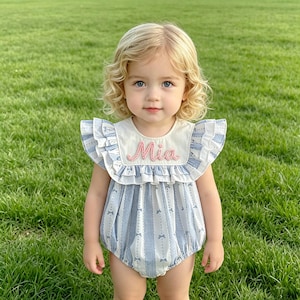 Puede incluir: Un body para beb&eacute; con un estampado de rayas azules y blancas y detalles de lazos. El body tiene un cuello color crema con el nombre "Mia" bordado en rosa. El body tiene mangas con volantes y est&aacute; hecho de una tela suave.