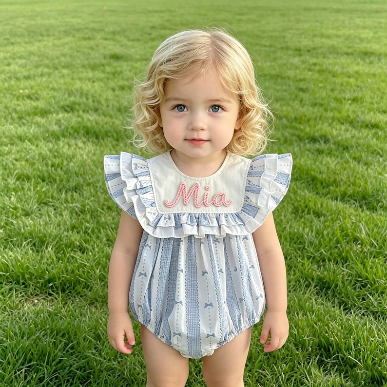 Puede incluir: Un body para beb&eacute; con un estampado de rayas azules y blancas y detalles de lazos. El body tiene un cuello color crema con el nombre "Mia" bordado en rosa. El body tiene mangas con volantes y est&aacute; hecho de una tela suave.