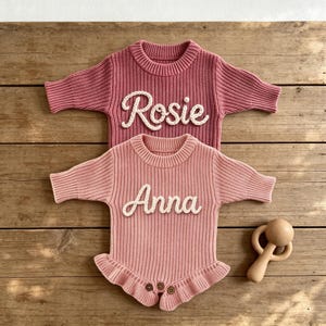 May include: Two long-sleeved knitted rompers in dusty rose and light pink. The top romper features the name Rosie in white script, while the bottom romper features the name Anna in white script. Both have ribbed textures and a wooden rattle.