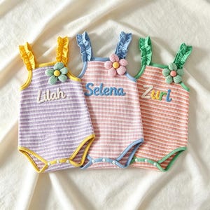 Puede incluir: Tres bodies de bebé con los nombres Lilah, Selena y Zuri bordados. Cada uno tiene un patrón de rayas en morado, rosa y naranja, con detalles de flores y tirantes con volantes. Los bodies tienen ribetes amarillos, azules y verdes.