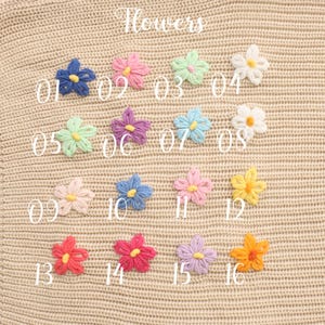 May include: A collection of sixteen small, crocheted flower appliques arranged in a grid on a beige knit fabric. Each flower features five petals in various colors including blue, pink, green, white, purple, yellow, and red, with contrasting centers.