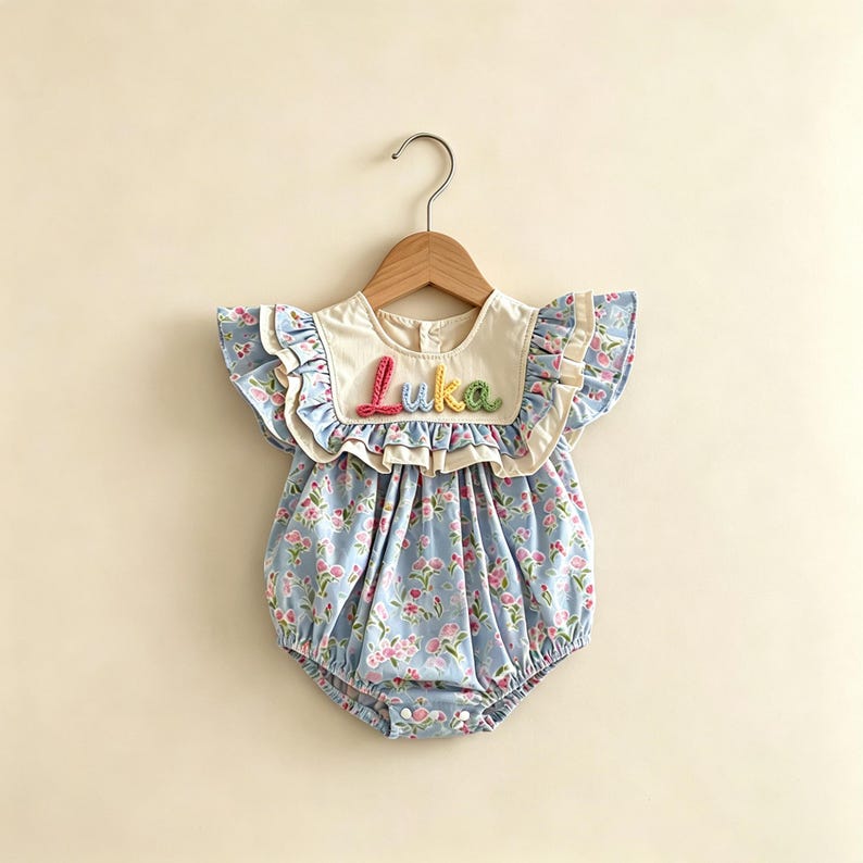 Puede incluir: Un pelele de beb&eacute; con estampado floral azul claro y mangas con volantes. El pelele tiene un cuello color crema con el nombre "Luka" bordado en hilo colorido. La prenda cuelga de una percha de madera.