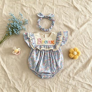 Puede incluir: Conjunto de beb&eacute; con ranita y diadema con estampado floral en azul claro y rosa. La ranita tiene un cuello color crema con el nombre "Bronx" bordado en letras coloridas. Incluye una diadema a juego con lazo.