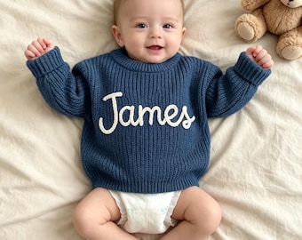 May include: A navy blue long-sleeved knit sweater featuring the name James embroidered in white script lettering across the chest. The sweater has a ribbed crew neckline, ribbed cuffs, and a ribbed hem, paired with a white disposable diaper.
