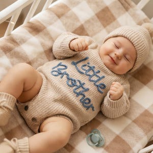May include: A beige knitted long-sleeve romper with the text "little brother" embroidered in blue yarn across the chest. The outfit includes a matching beige knit beanie and knit booties. A light blue pacifier rests nearby on the soft fabric.