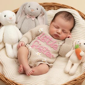 May include: A sleeping infant wearing a beige long-sleeved knit romper with the pink embroidered text little sister and small white flower accents. The romper features two wooden buttons at the bottom. Three plush toys, including two rabbits and a bear, surround the infant inside a woven basket.