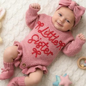 May include: A dusty pink knitted long-sleeve romper featuring the red embroidered text "little sister" and small bow accents. The garment includes ruffled leg openings, three small wooden buttons at the crotch, matching knitted booties, and a headband.