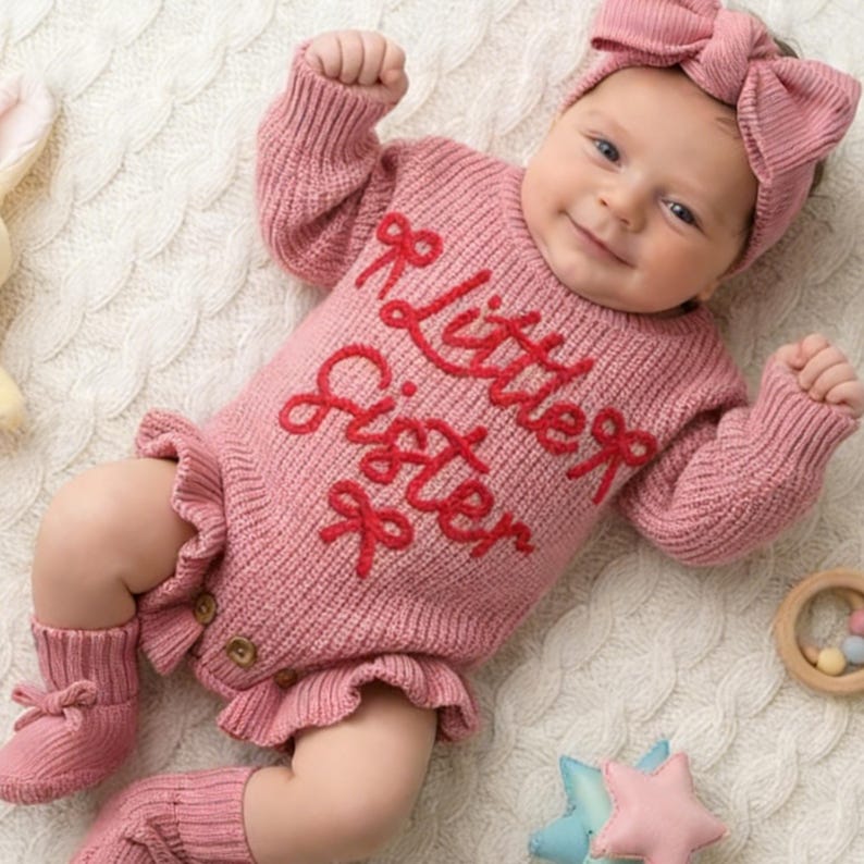 May include: A dusty pink knitted long-sleeve romper featuring the red embroidered text "little sister" and small bow accents. The garment includes ruffled leg openings, three small wooden buttons at the crotch, matching knitted booties, and a headband.