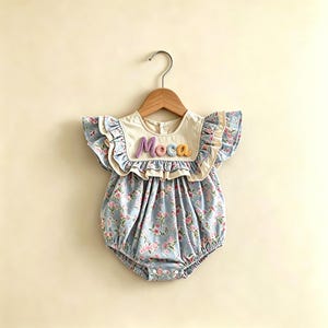 Puede incluir: Un body para beb&eacute; con estampado floral en azul claro y rosa. El body tiene un cuello color crema con el nombre "Moca" bordado en hilo colorido. Mangas con volantes y una percha de madera completan el look.