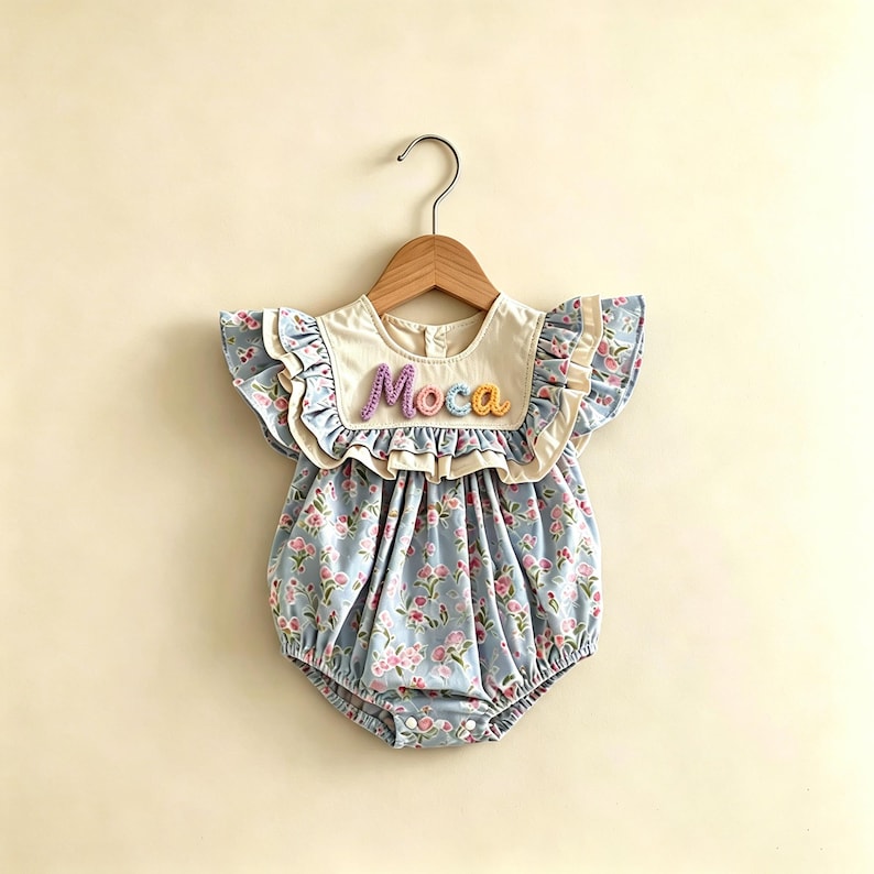 Puede incluir: Un body para beb&eacute; con estampado floral en azul claro y rosa. El body tiene un cuello color crema con el nombre "Moca" bordado en hilo colorido. Mangas con volantes y una percha de madera completan el look.