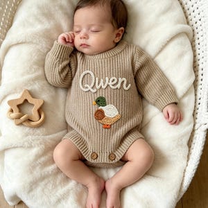 Puede incluir: Un pelele de punto acanalado en color beige de manga larga con el nombre Qwen bordado en letra blanca y una pequeña aplicación de un pato real con detalles en verde, blanco y marrón. La prenda incluye dos pequeños botones marrones en el bajo.
