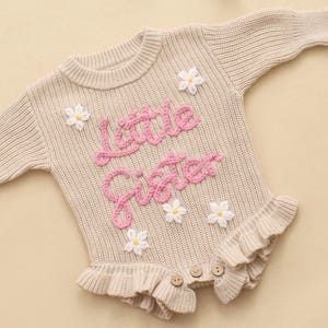 May include: A beige long-sleeved knit romper featuring the pink embroidered text "little sister" across the chest. The garment is decorated with several small white daisy appliqu&eacute;s with yellow centers and includes a ruffled hem with three buttons.