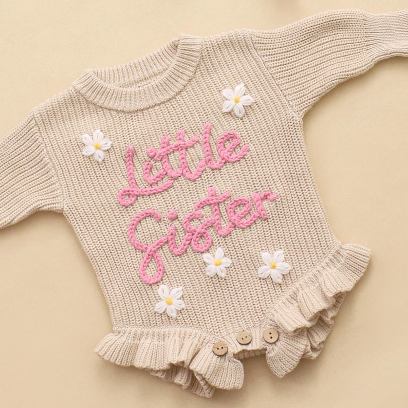 May include: A beige long-sleeved knit romper featuring the pink embroidered text "little sister" across the chest. The garment is decorated with several small white daisy appliqu&eacute;s with yellow centers and includes a ruffled hem with three buttons.