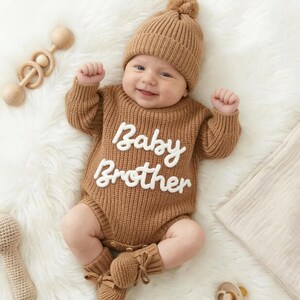 May include: A tan knitted baby romper featuring long sleeves, a ribbed texture, and the white embroidered text "Baby Brother" across the chest. The outfit includes a matching tan knitted beanie with a pom-pom and tan knitted booties with ties.