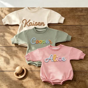 Könnte beinhalten: Drei langärmelige Baby-Strampler in Creme, Salbeigrün und Altrosa. Jeder Strampler hat einen Rundhalsausschnitt und einen personalisierten Namen, der in einer erhabenen, strukturierten Garnschrift gestickt ist: Kaisen, Cooper und Alice.