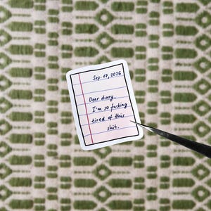Op de afbeelding: Een sticker die lijkt op een gelijnde notitiepagina met de tekst "Sep. 17, 2026" en "Dear diary, I'm so fucking tired of this shit." De sticker wordt vastgehouden door een pincet tegen een groen-witte achtergrond met patroon.