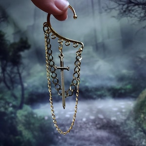 May include: Gold-toned ear cuff featuring a sword pendant and chain accents. The ear cuff has a curved design that hooks over the ear. The sword pendant hangs from a chain, adding a touch of medieval flair. The background is a blurred forest scene.