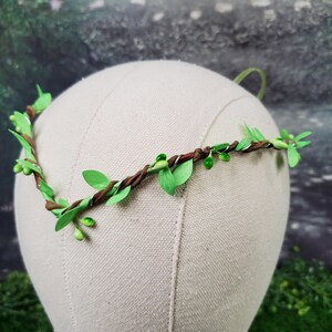May include: A handcrafted floral crown featuring a brown vine base adorned with green leaves and small green berries. The crown is displayed on a mannequin head, showcasing its delicate design. Perfect for weddings, festivals, or themed events.