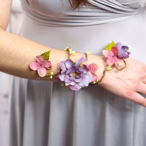 May include: A floral wrist cuff featuring pink and purple fabric flowers and green leaves. The flowers are attached to a thin, brown wire base. The cuff is worn on an arm, with a light gray dress in the background.
