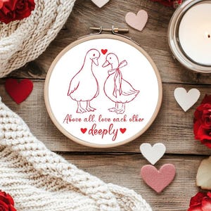 May include: A wooden embroidery hoop with a white fabric featuring a red line drawing of two ducks facing each other. The text "Above all, love each other deeply" is below the ducks. Red and pink hearts and roses surround the hoop.