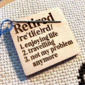 May include: A square wooden tag with the word "Retired" and its phonetic pronunciation. The tag lists "enjoying life," "travelling," and "not my problem anymore." A needle and blue thread are attached.