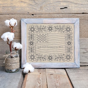 May include: A framed artwork with an American flag design and floral border. The flag has stars and stripes, with the text "God shed his grace on thee." The frame is light grey, and the artwork is displayed on a wooden surface.