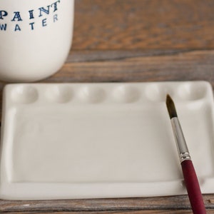May include: A white ceramic paint palette with six wells and a rectangular mixing area. A paintbrush with a silver ferrule and a burgundy handle rests on the palette. A white ceramic container with the words "PAINT WATER" in blue lettering is in the background.