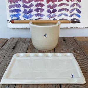 May include: A cream ceramic paint cup with a blue "J" and a matching rectangular palette. A paintbrush rests on the cup. A watercolour chart with floral shapes is in the background. The items are on a wooden surface.