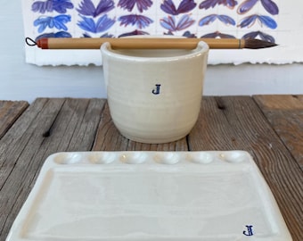 May include: A cream-colored ceramic paint cup with a blue "J" and a matching rectangular palette. A paintbrush rests on the cup. A watercolor chart with floral shapes is in the background. The items are on a wooden surface.