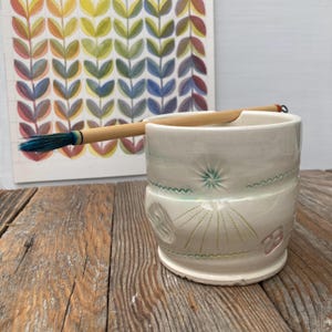 May include: A handmade ceramic cup with a light beige glaze, featuring embossed designs and a turquoise starburst. A wooden paintbrush with a blue tip rests inside. A colorful floral painting is in the background.