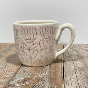 Puede incluir: Taza de cerámica color crema con un diseño floral en rosa. Las flores están estilizadas y se repiten alrededor de la taza. Tiene un asa redondeada y está sobre una superficie de madera.