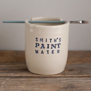 May include: Cream-colored ceramic paint water container with the text "SMITH'S PAINT WATER" in dark blue. A light blue paintbrush and a paint brush are resting on the rim of the container. The container is sitting on a wooden surface.